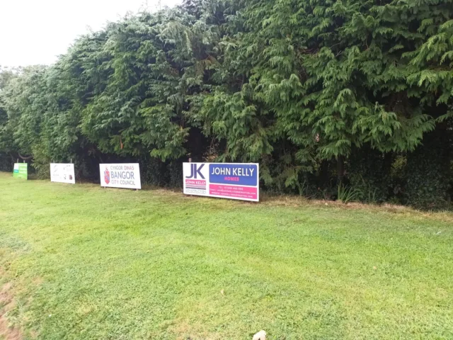 A photograph showing a John Kelly Construction and John Kelly Homes sponsorship board on the Bangor Cricket Club grounds next to some other sponsorship boards.
