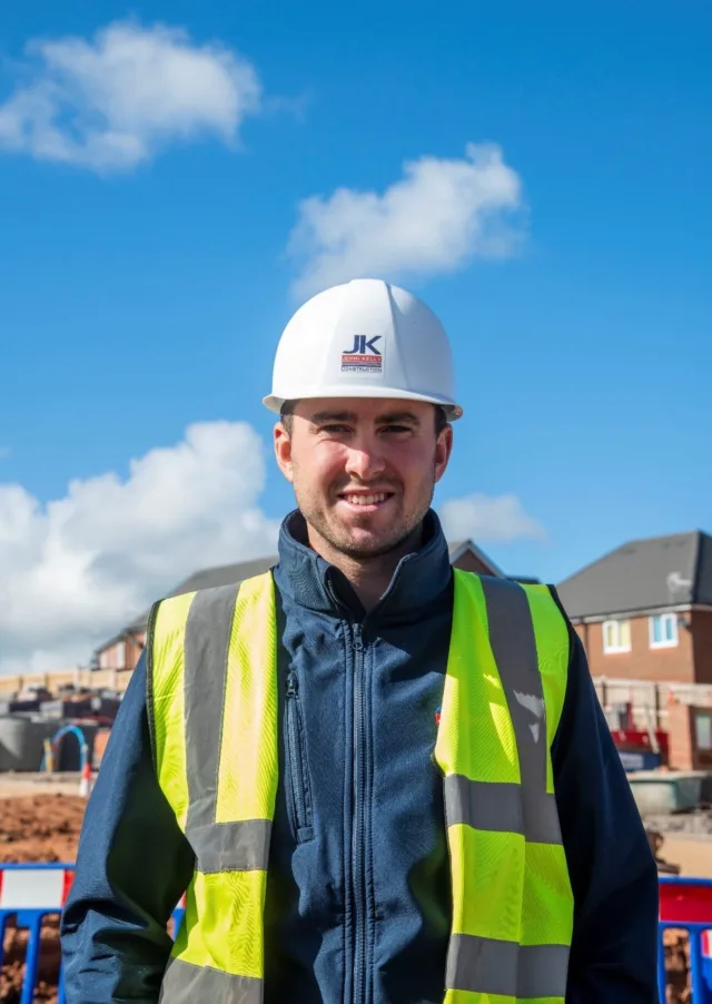 A photograph of our Site Manager Rhun smiling at the camera and wearing a white hard hat with a high-vis vest on.